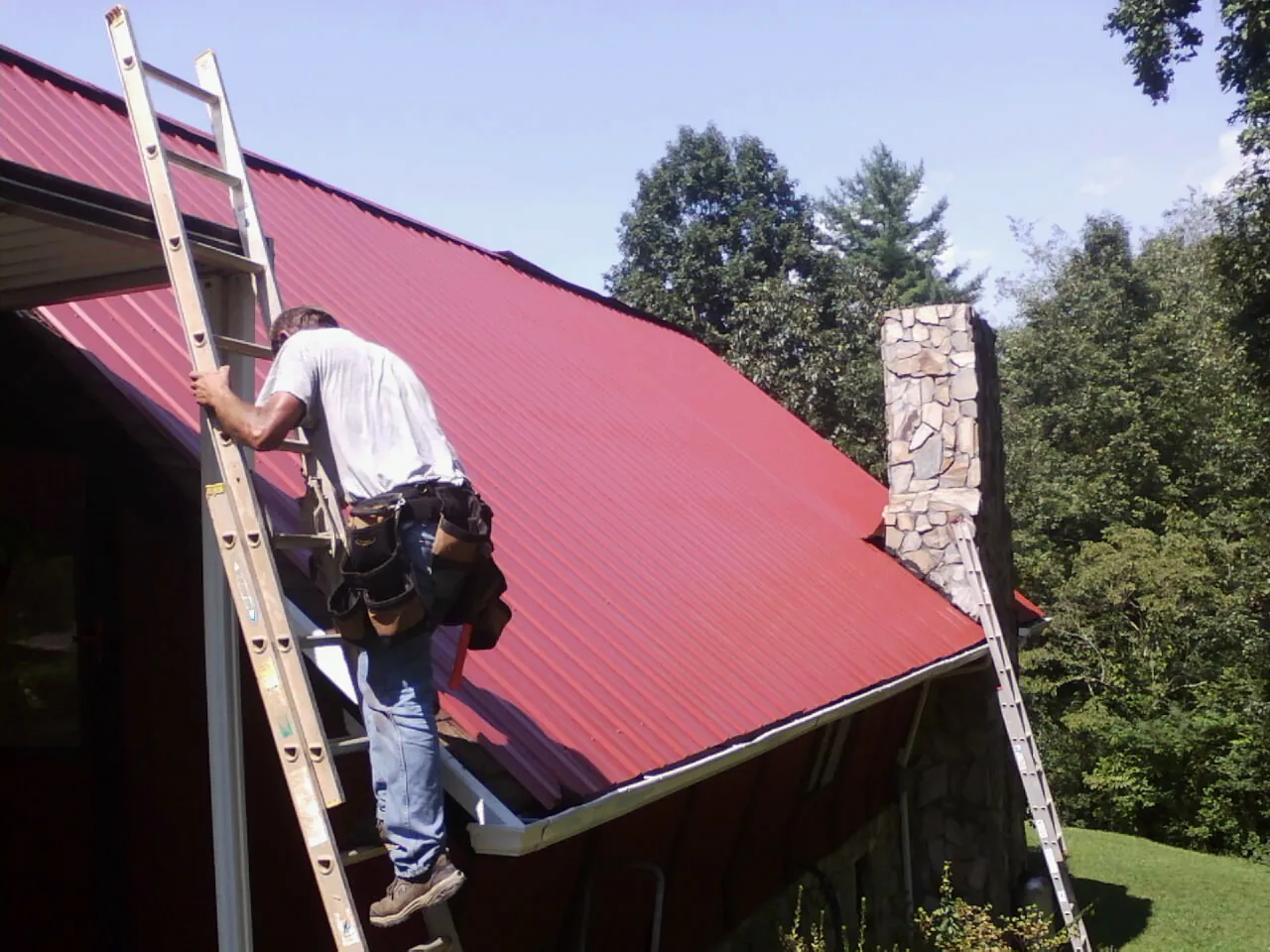 Roofer on a ladder beside a red metal roof and stone chimney — Soffit & Fascia Repair by Roofing Brothers in Campti